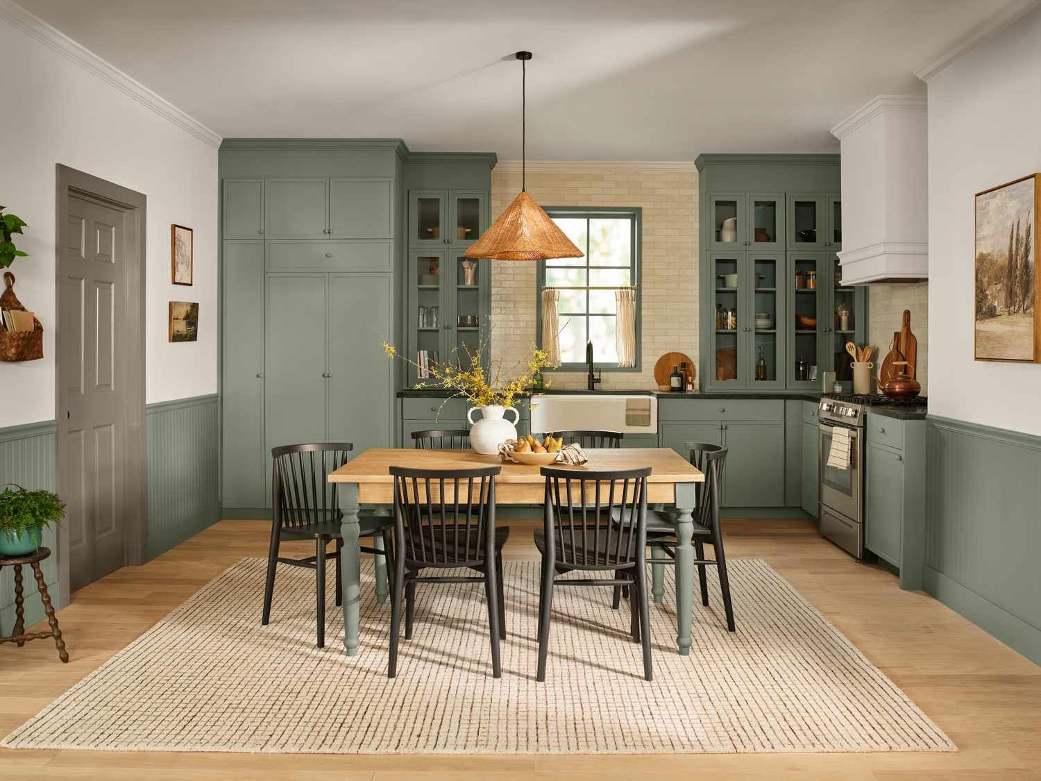 Dining room with sage green cabinets, wooden table, black chairs, and woven rug.