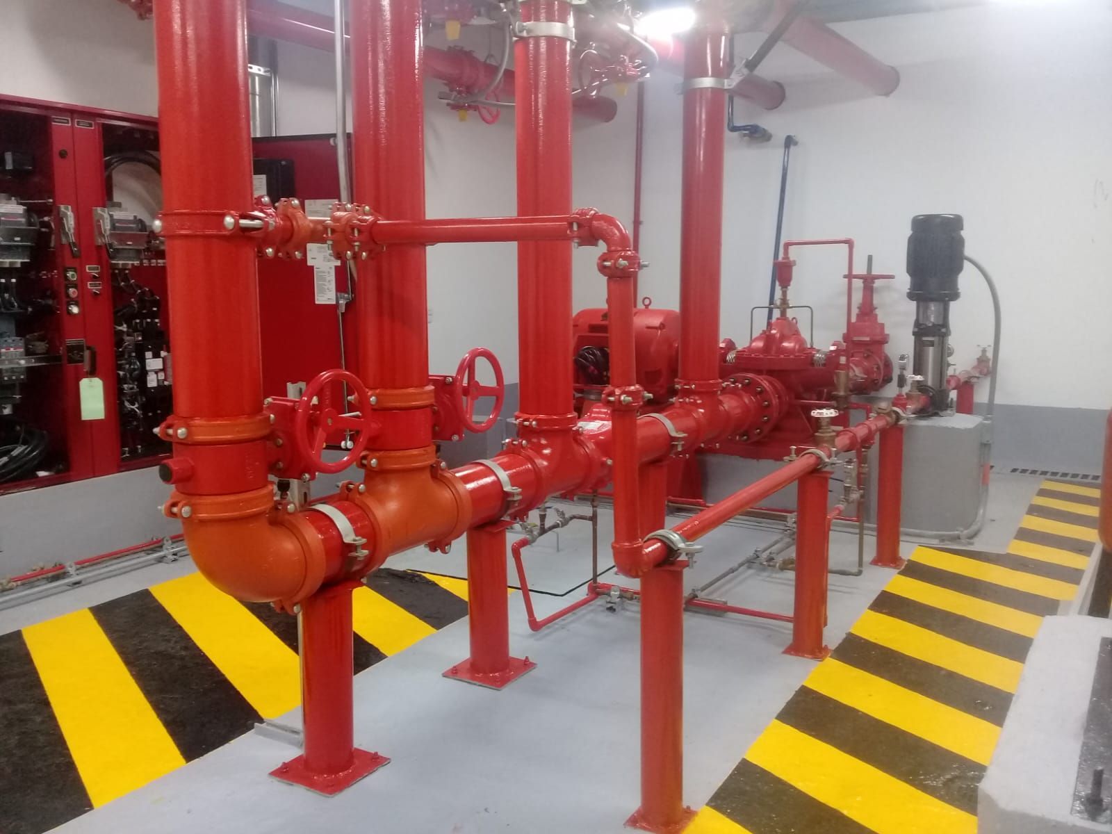 Tuberías y equipos de un sistema de extinción de incendios de color rojo en un cuarto de servicio, con marcas de peligro en el piso de color amarillo y negro.