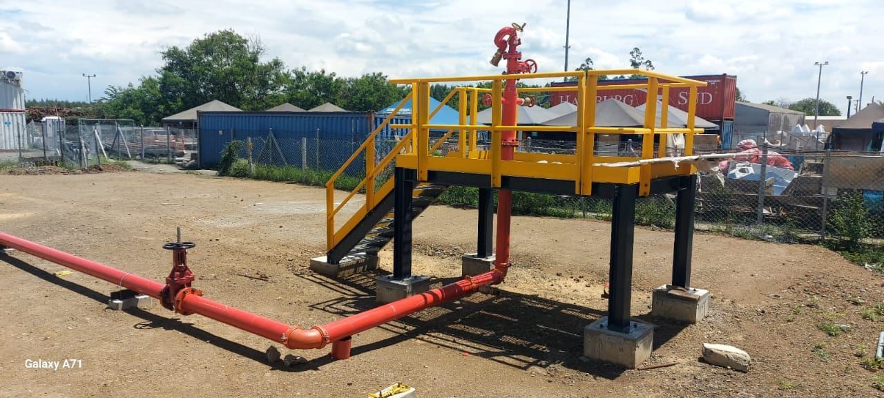 Plataforma elevada de color amarillo y negro con ribetes rojos y escalera, al aire libre sobre tierra.