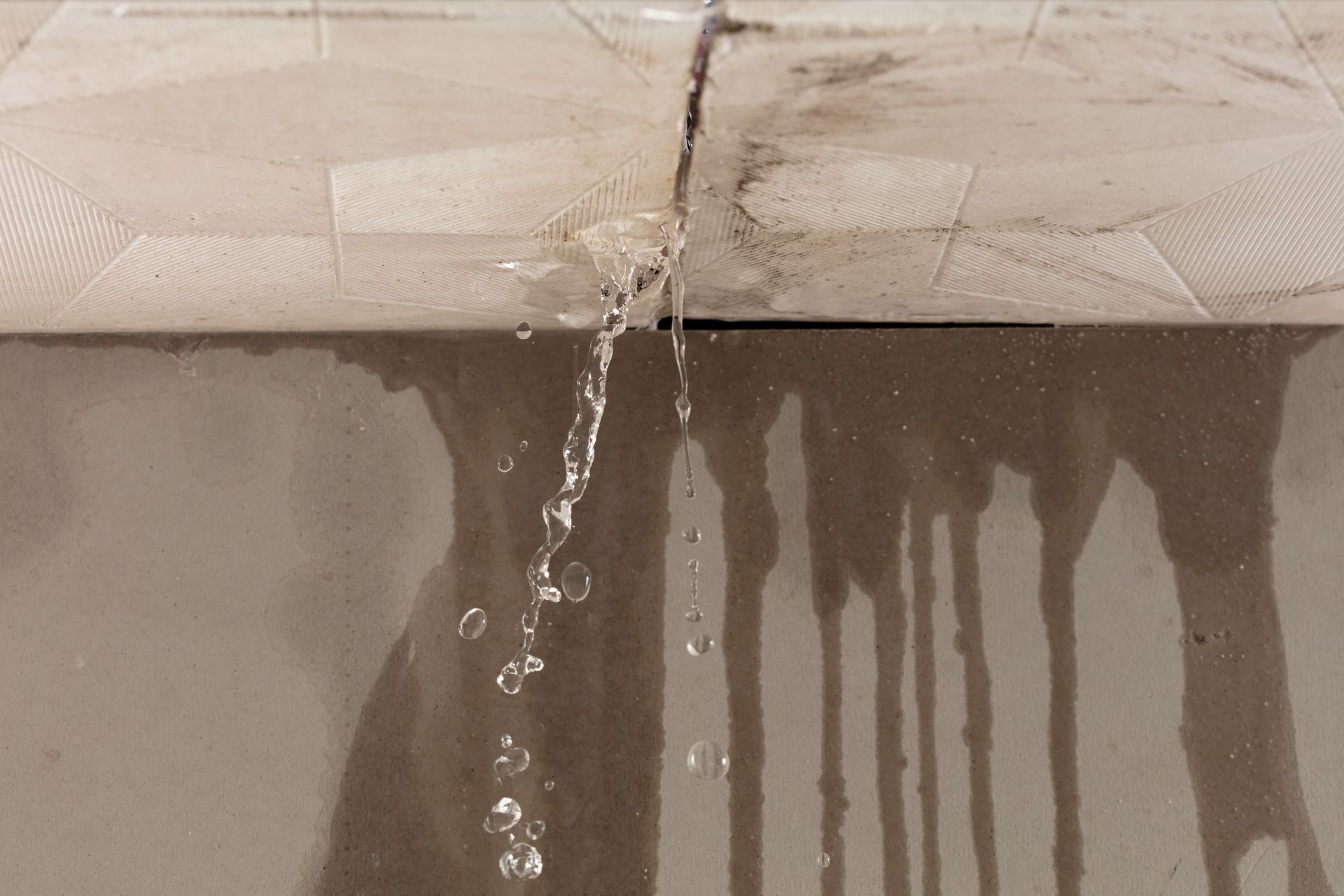 Water leaks through a crack between two light-colored, grimy tiles onto a smooth, reflective brown surface below.