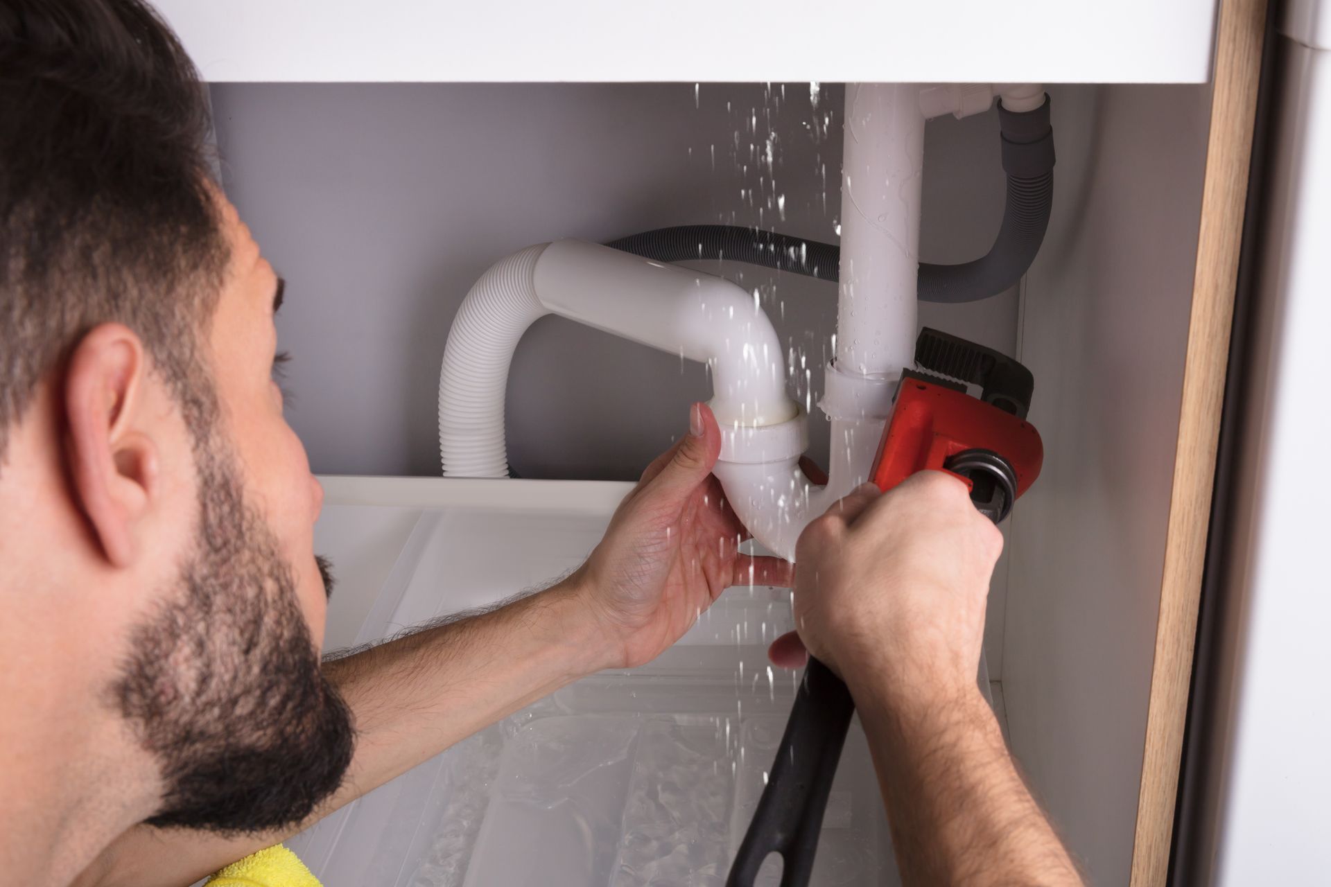 A person uses a red pipe wrench to repair a leaking white PVC pipe joint under a sink.