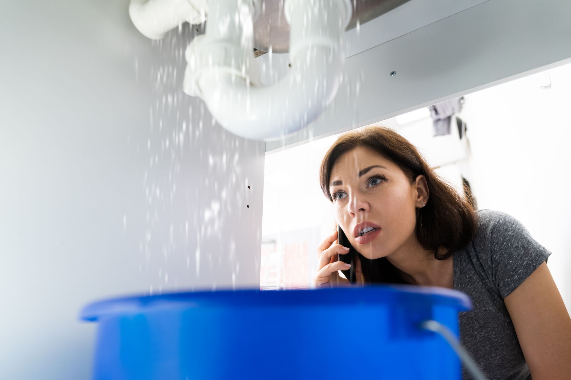 A person looks worried while talking on a phone, watching water leak from under-sink pipes into a blue bucket.