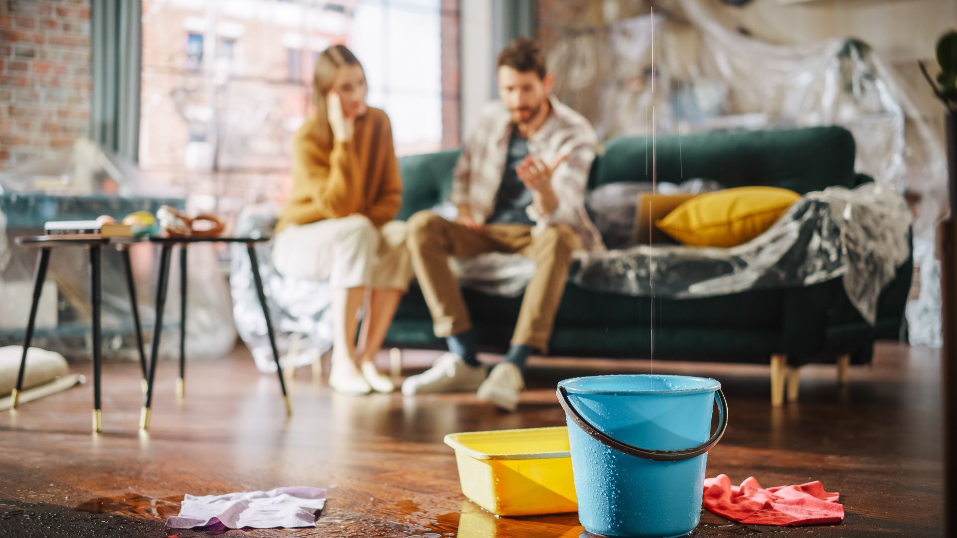 A couple sits on a sofa in a living room, looking concerned as a blue bucket catches water leaking from the ceiling.