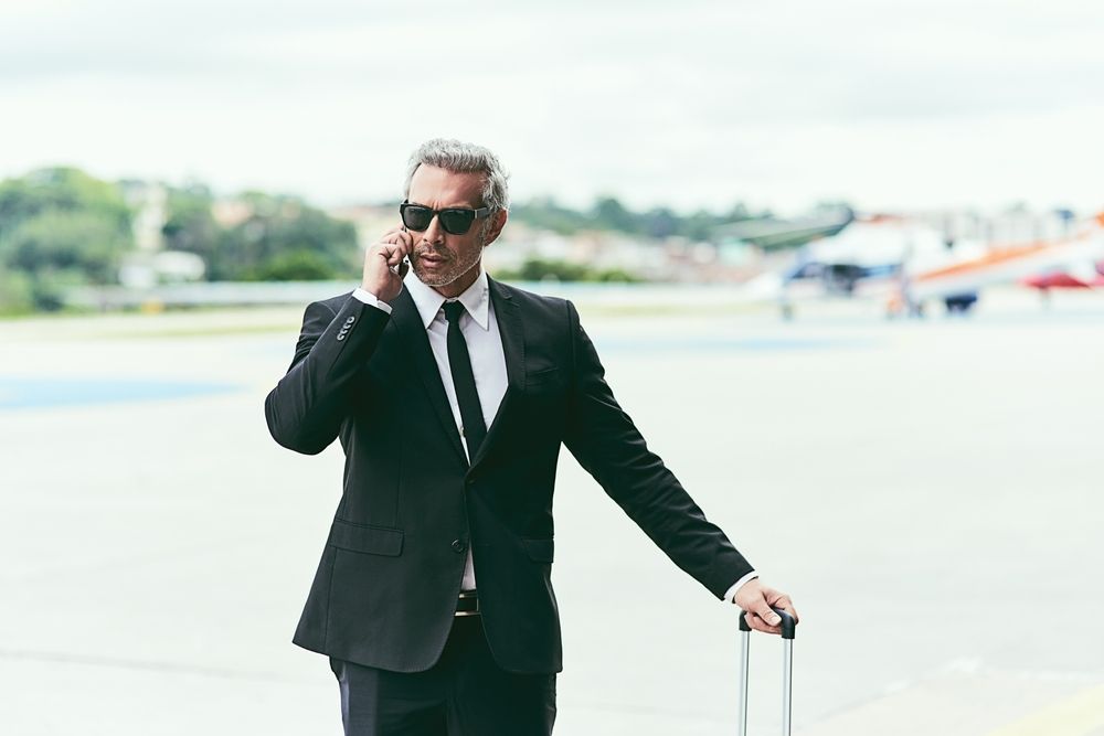 A man in a suit is talking on a cell phone while holding a suitcase.