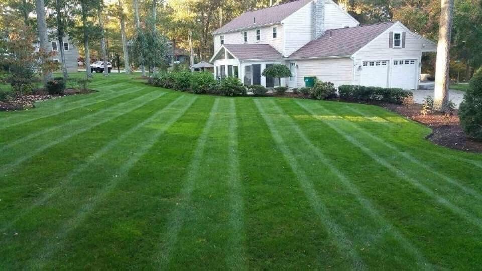 A two-story, light-colored house with a front porch and a neatly mowed, striped lawn featuring mulched landscaping beds.