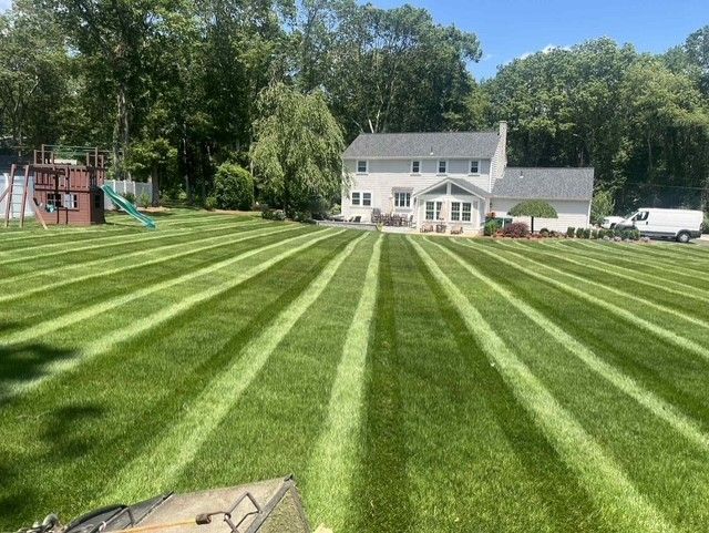 A person kneeling in a garden bed, spreading mulch in front of a house with a white wraparound porch and green lawn.
