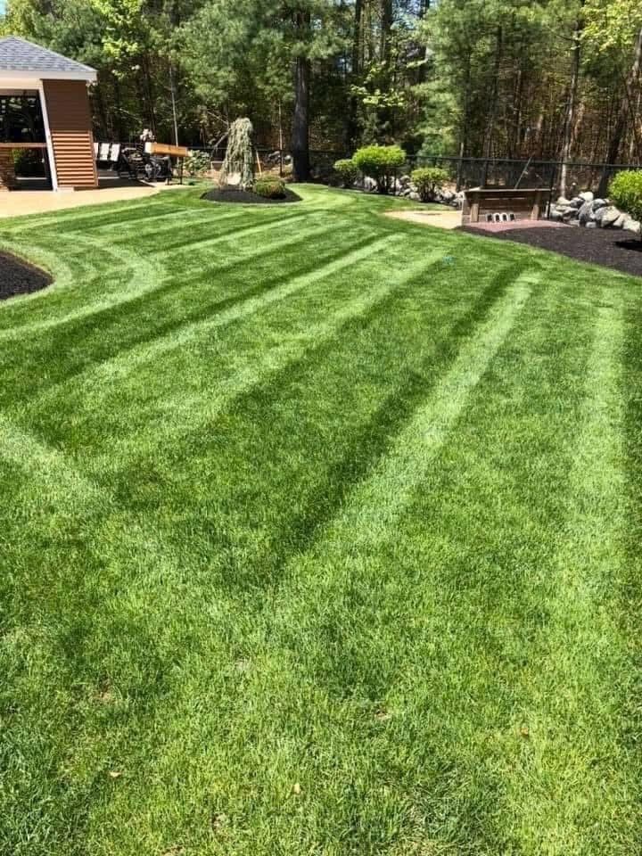 A person on a yellow riding lawnmower cuts green grass in front of a white barn-style building.