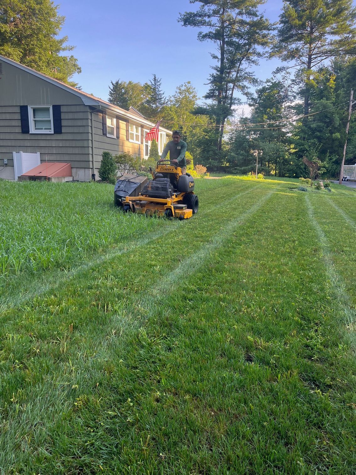 A person on a yellow zero-turn lawn mower cuts grass in a suburban yard next to a tan house.