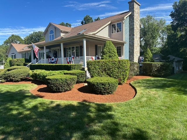 A tan two-story house with a stone chimney and wraparound porch, featuring manicured shrubs and red mulch garden beds.