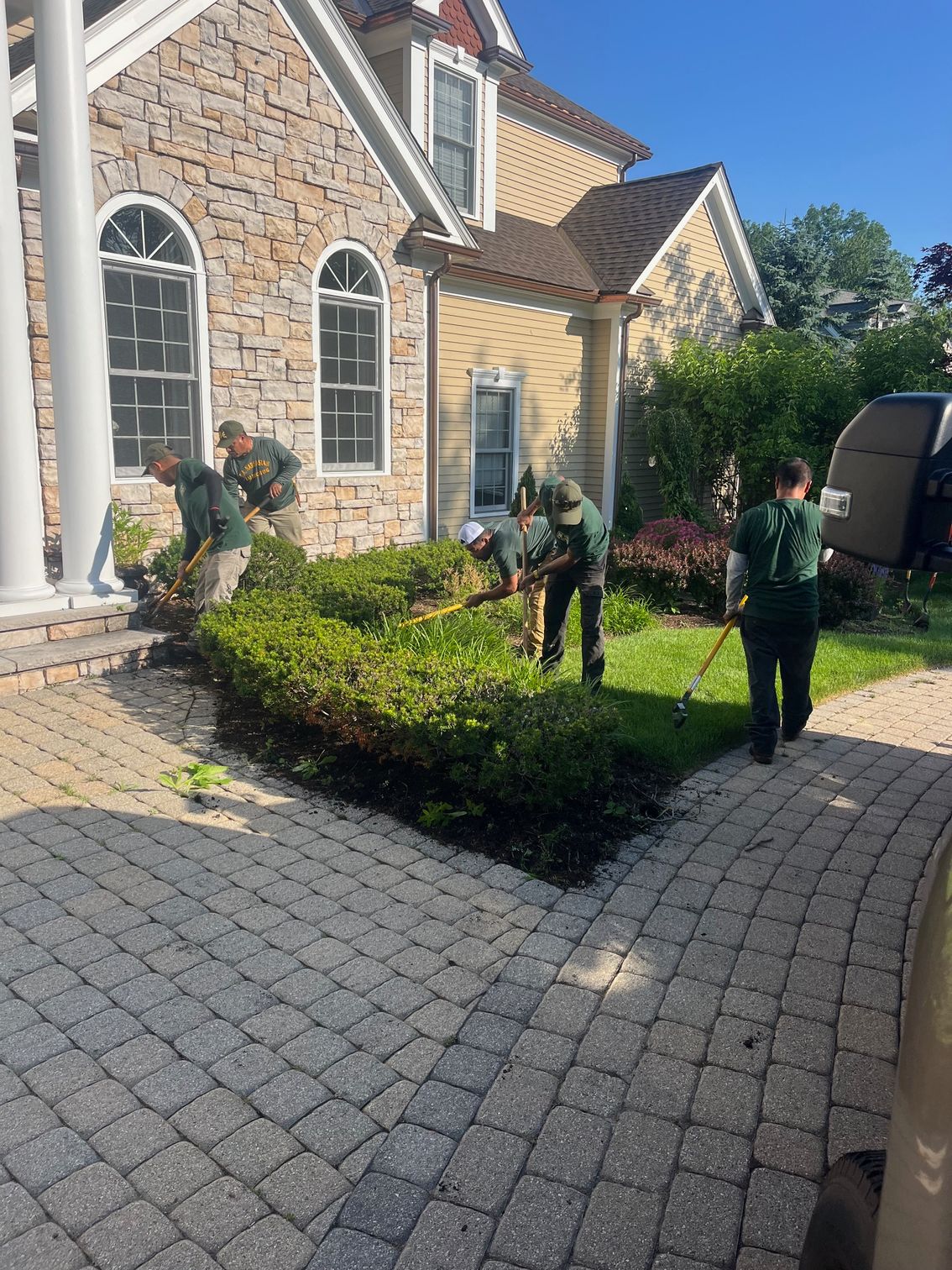 Landscapers in matching uniforms trim hedges and clear a lawn in front of a brick and stone house on a sunny day.