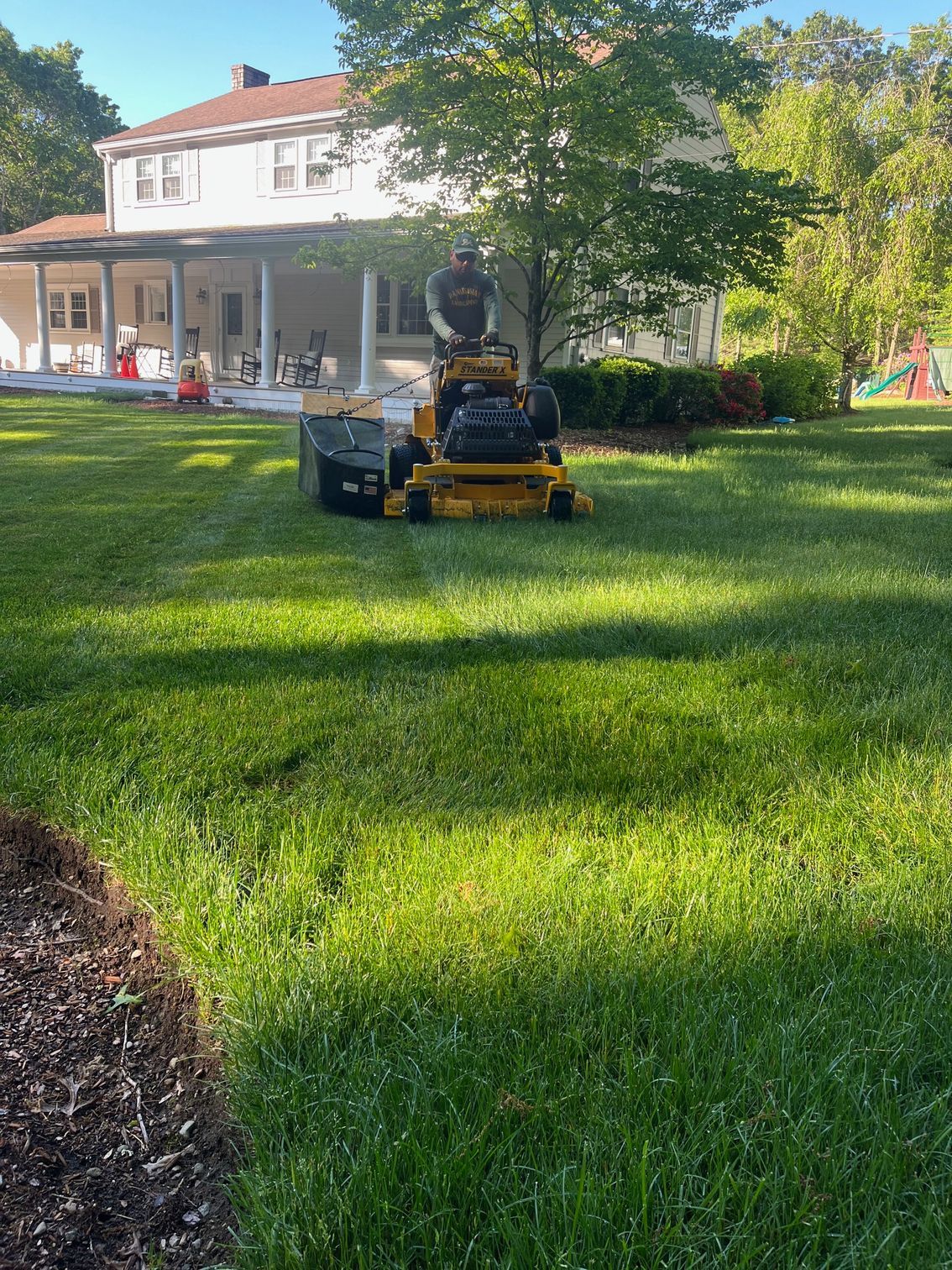 A person rides a yellow zero-turn mower across a sunny residential lawn in front of a white two-story house.