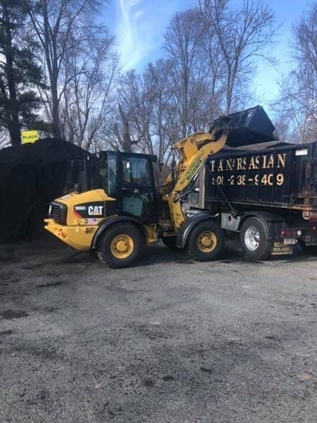 A yellow CAT wheel loader dumps soil into a black Tanurjian dump truck parked on a gravel lot under a clear sky.