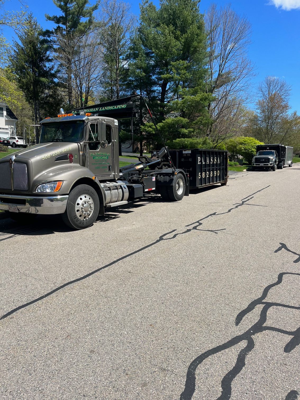 A brown roll-off truck hauling a black dumpster container parked on an asphalt road with a pickup truck in the distance.