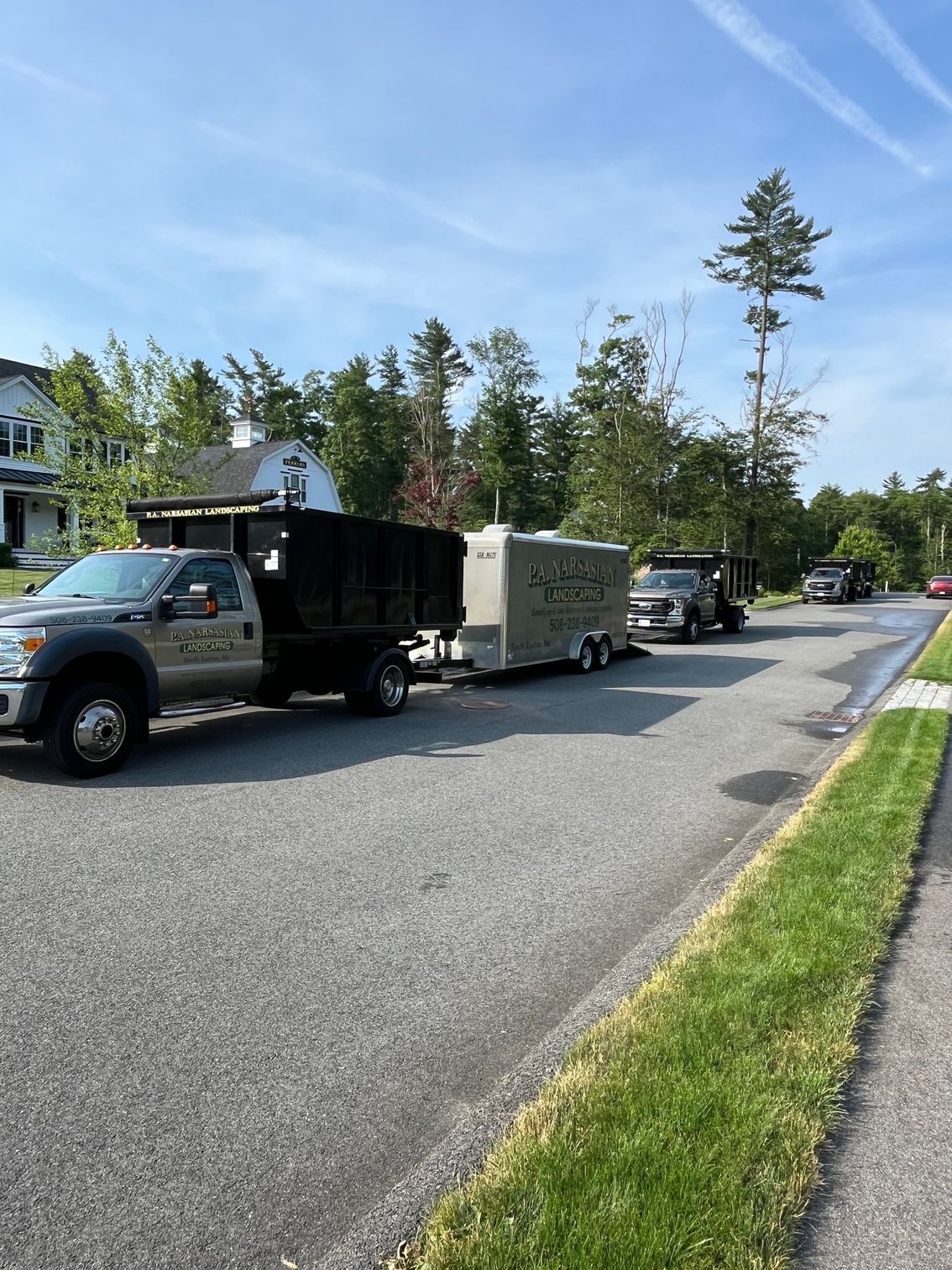 A dump truck and trailer parked on a sunny suburban street, with trees and a house in the background.