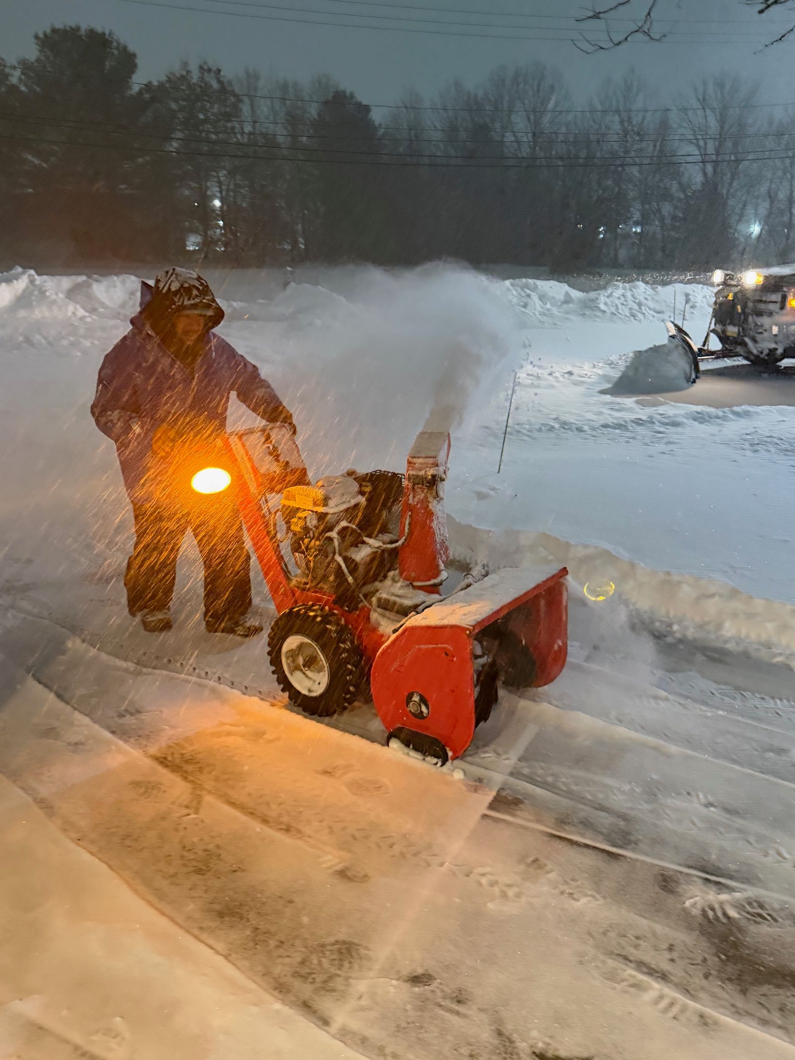 A yellow front-end loader moves a large pile of snow at night under bright work lights.