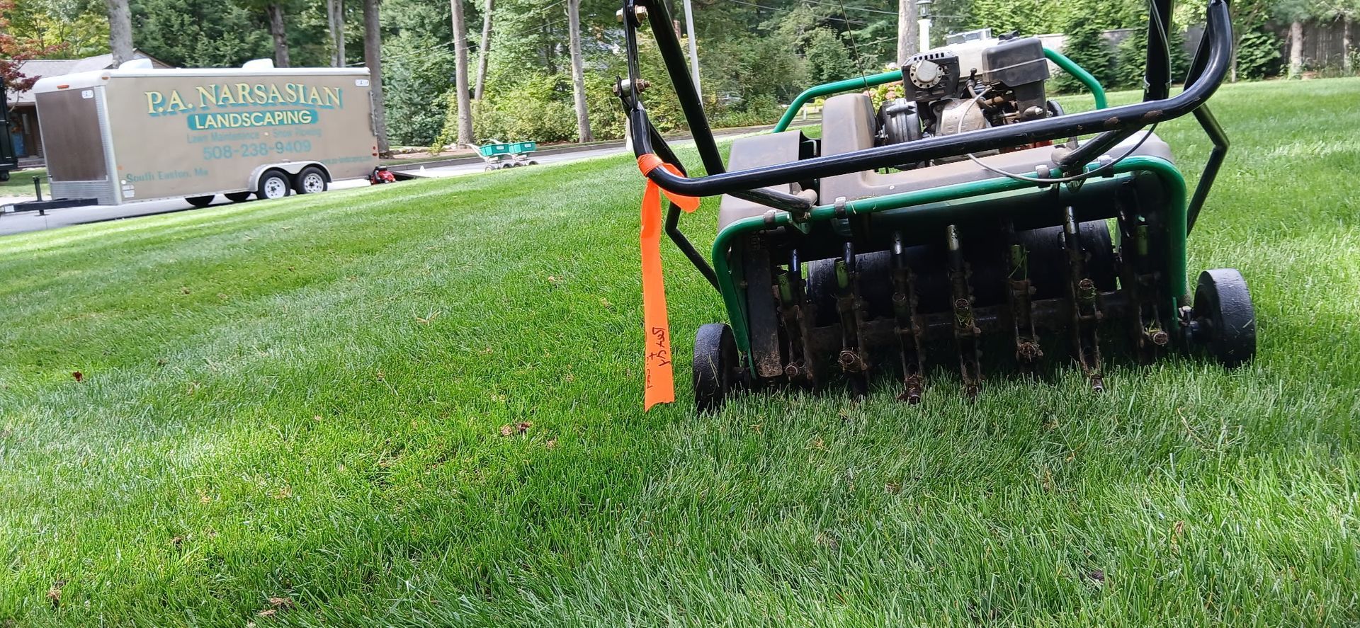 A green walk-behind lawn aerator sits on a mowed lawn with an orange flag nearby and a service trailer in the background.