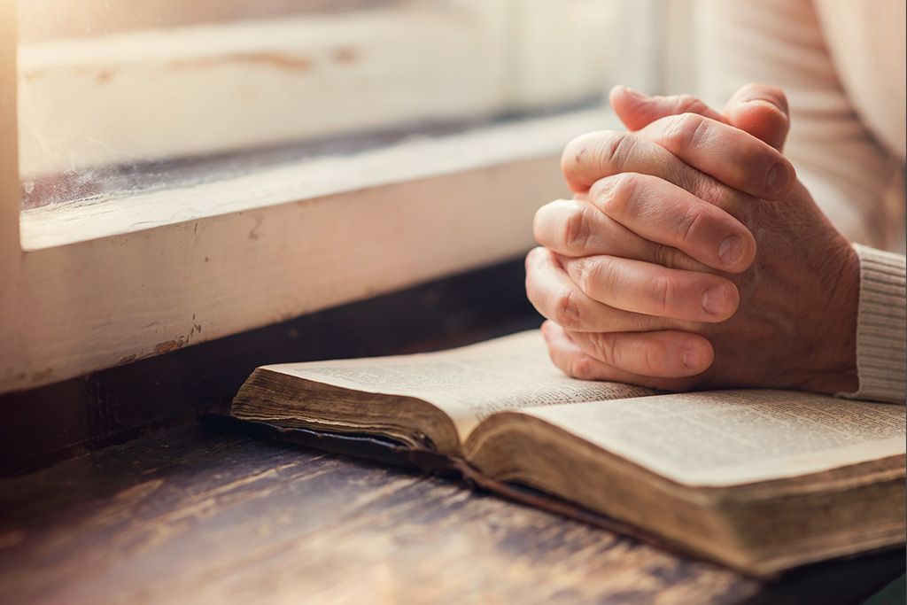 A person is praying with their hands folded over an open bible.