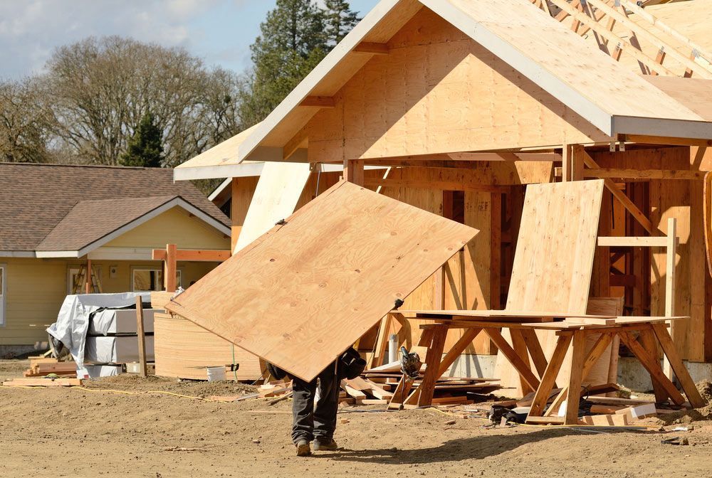 A Man Is Carrying A Piece Of Plywood In Front Of A House Under Construction — Trussted Frames & Trusses in Port Stephens, NSW