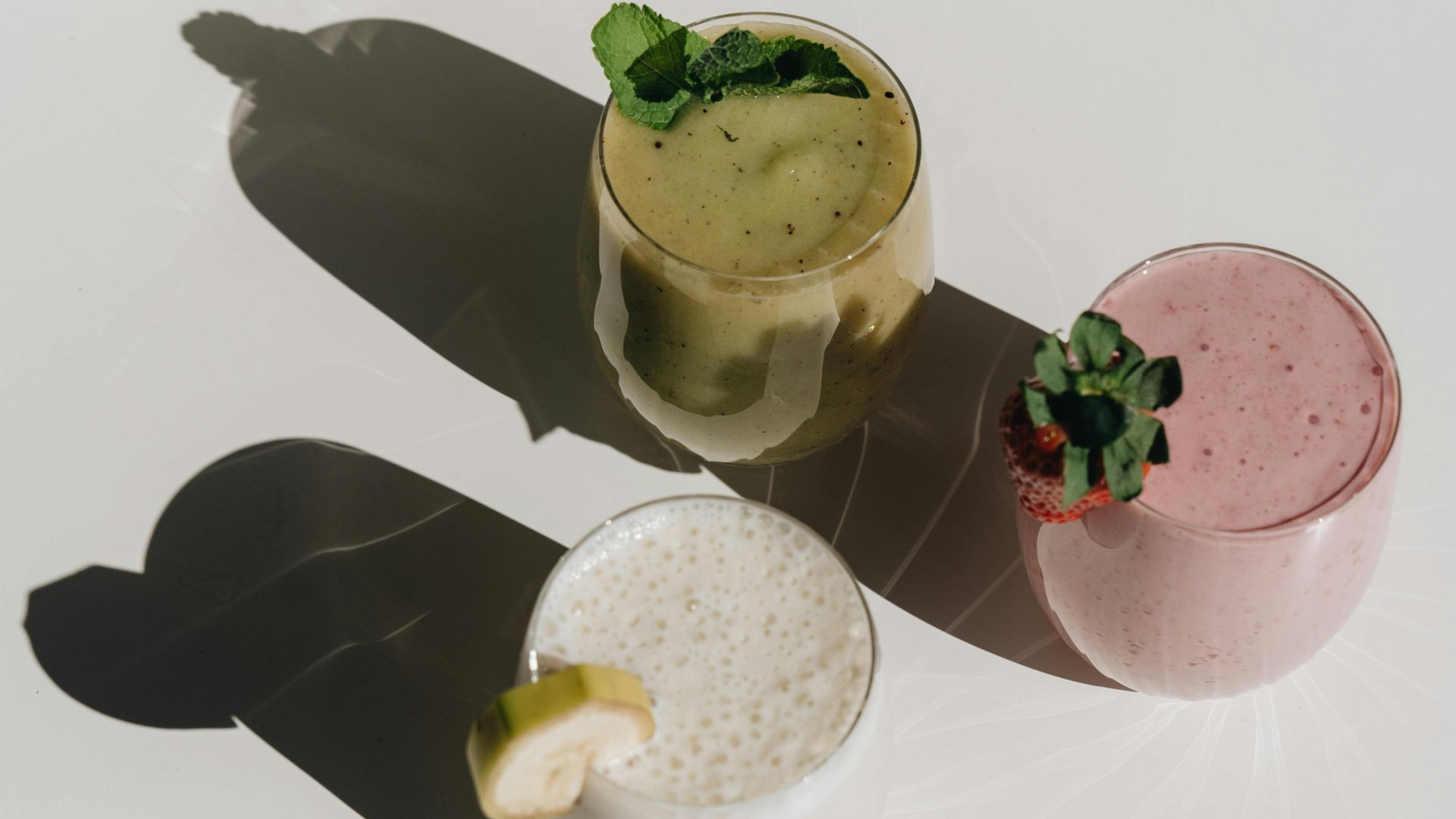 Three pastel cocktails with fruit garnish casting long shadows on a white surface.
