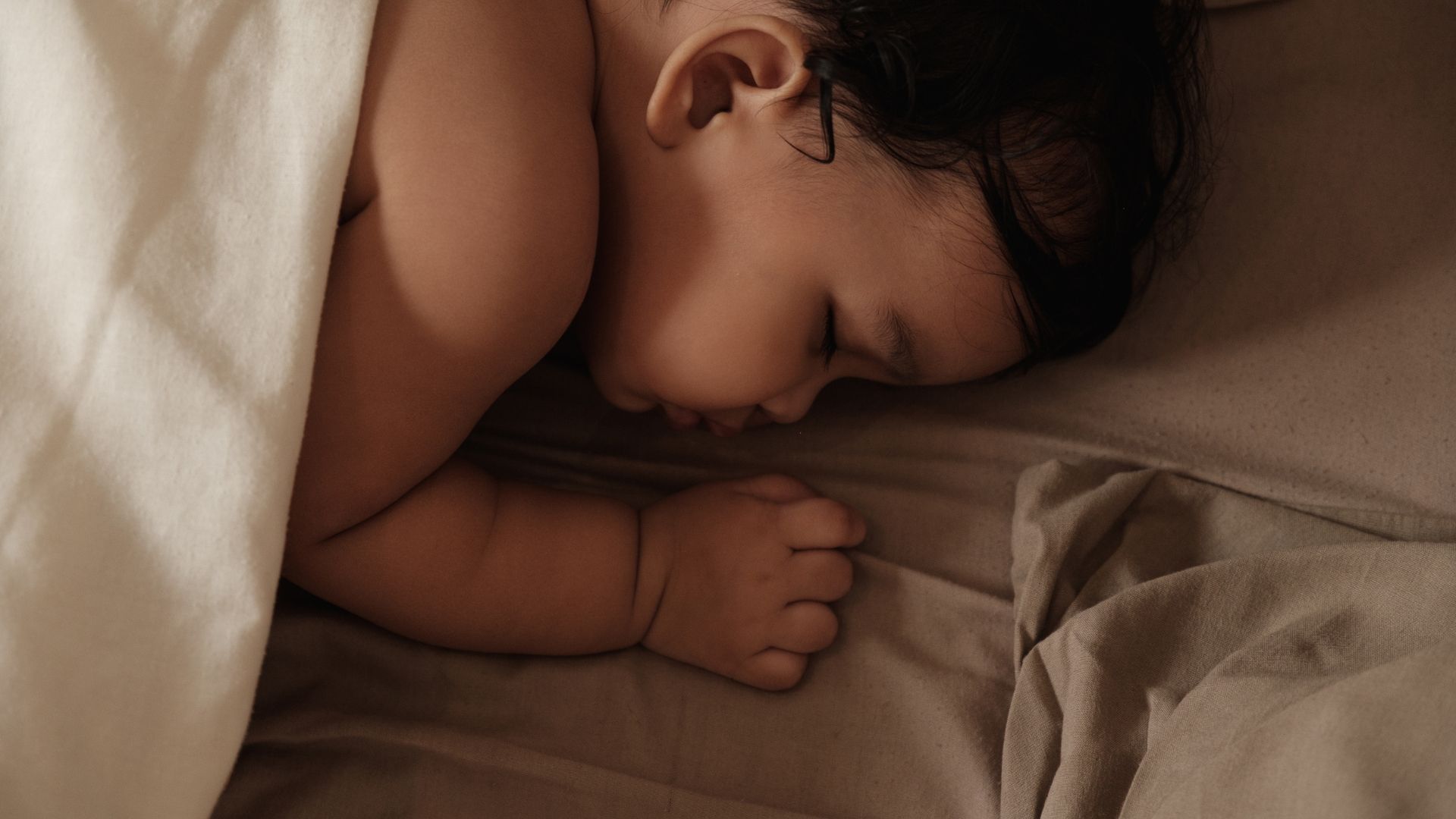 Baby asleep on a bed, covered by a sheet. Brown hair, skin, and bedding.