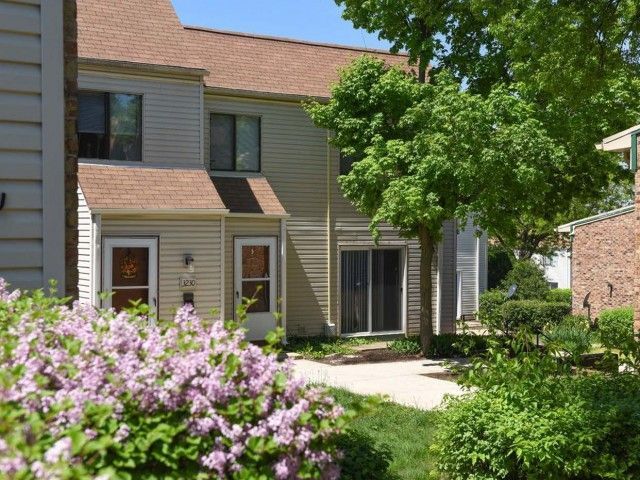 Townhouse with tan siding, front garden, and blooming pink shrubs beside a walkway under a leafy tree