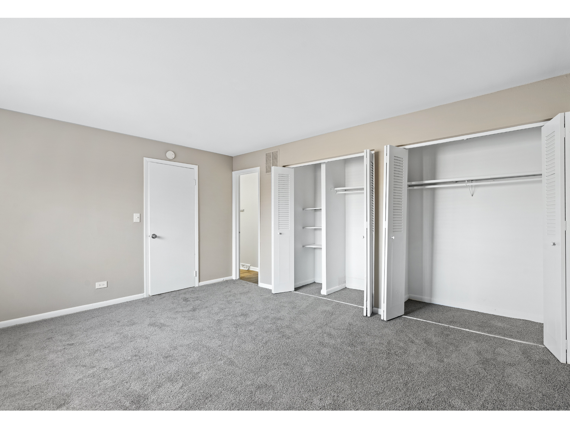 Empty beige bedroom with gray carpet, white closet doors, and a white door near a mirror.
