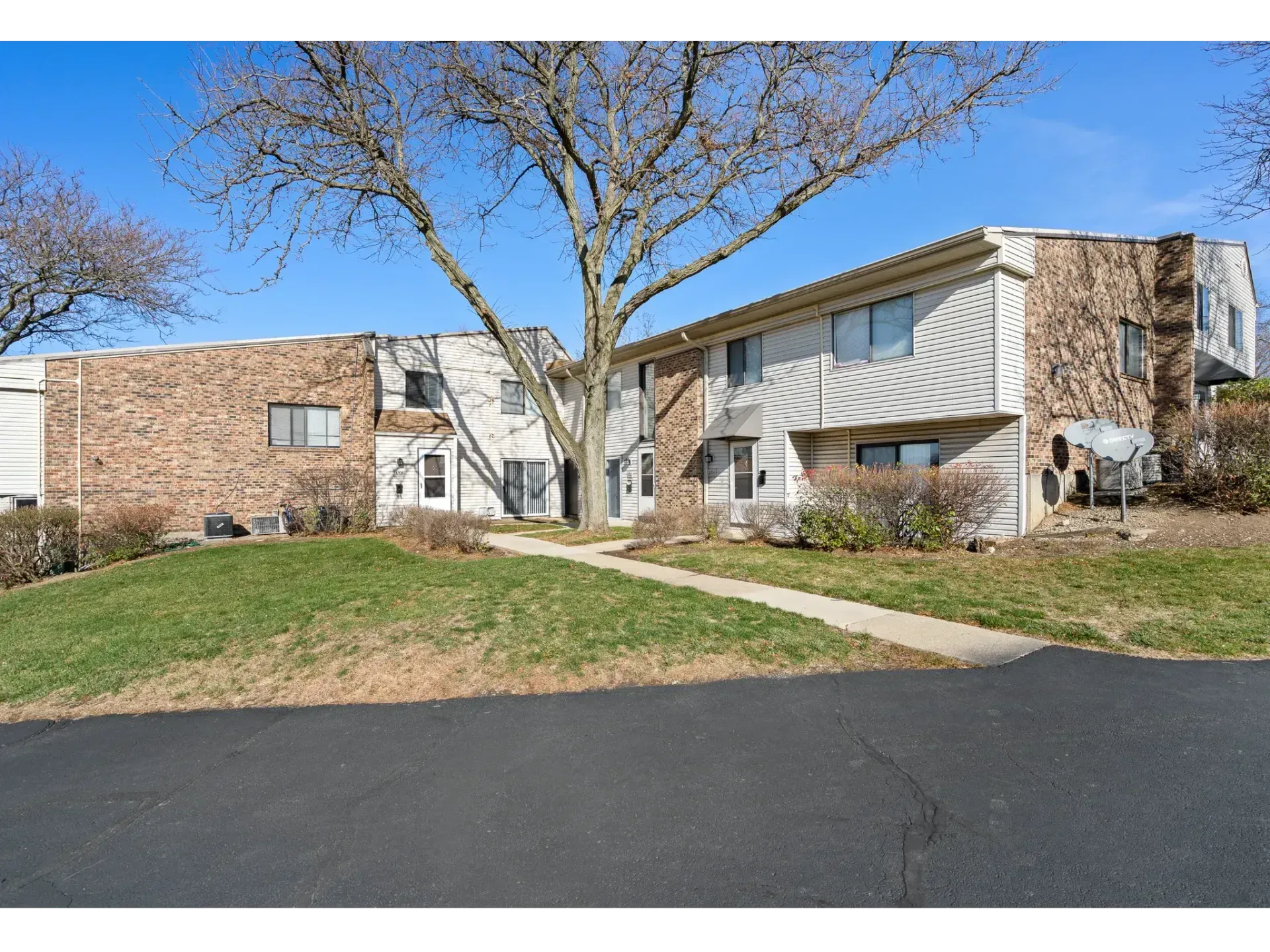 Apartment complex with brick and beige buildings, a tree, sidewalk, and empty parking area under a blue sky