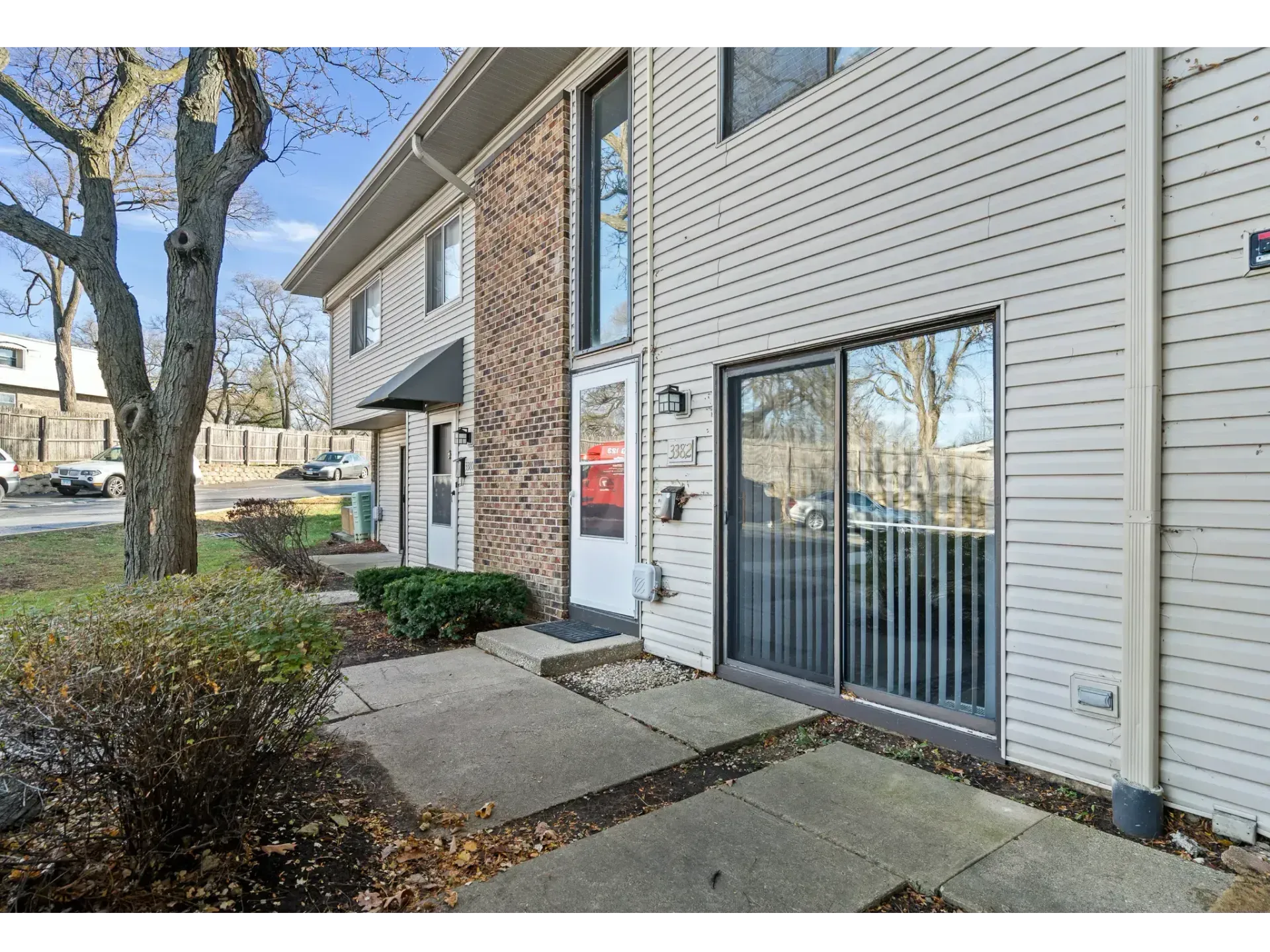 Apartment entrance with glass door, white siding, and concrete walkway beside trees and shrubs