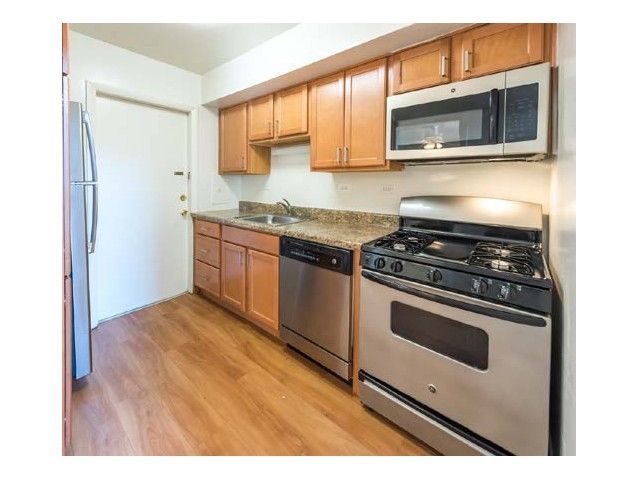 Compact kitchen with stainless appliances, wood cabinets, white walls, and light wood flooring.
