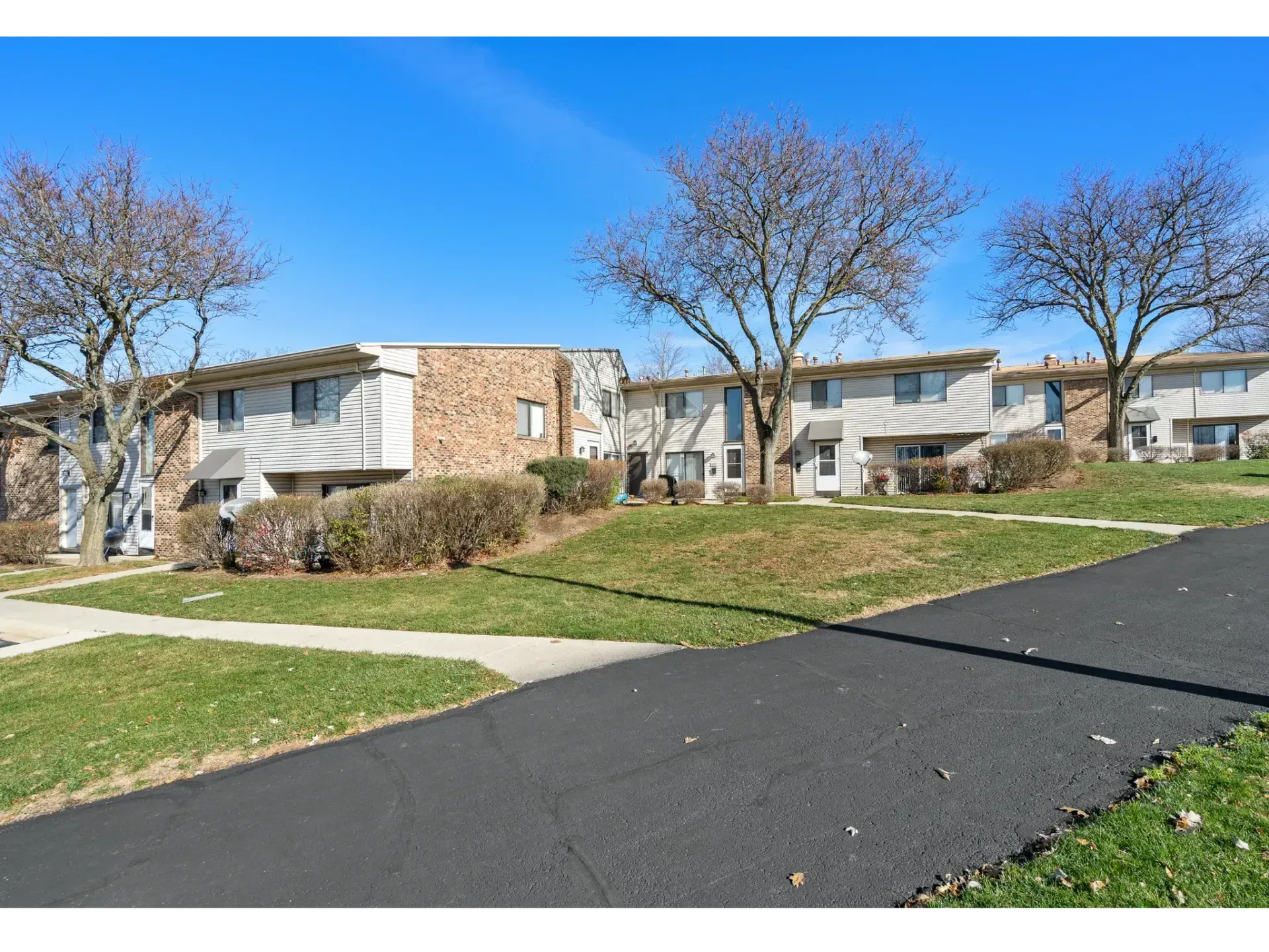 Apartment buildings beside a paved road under a bright blue sky with leafless trees and green lawns