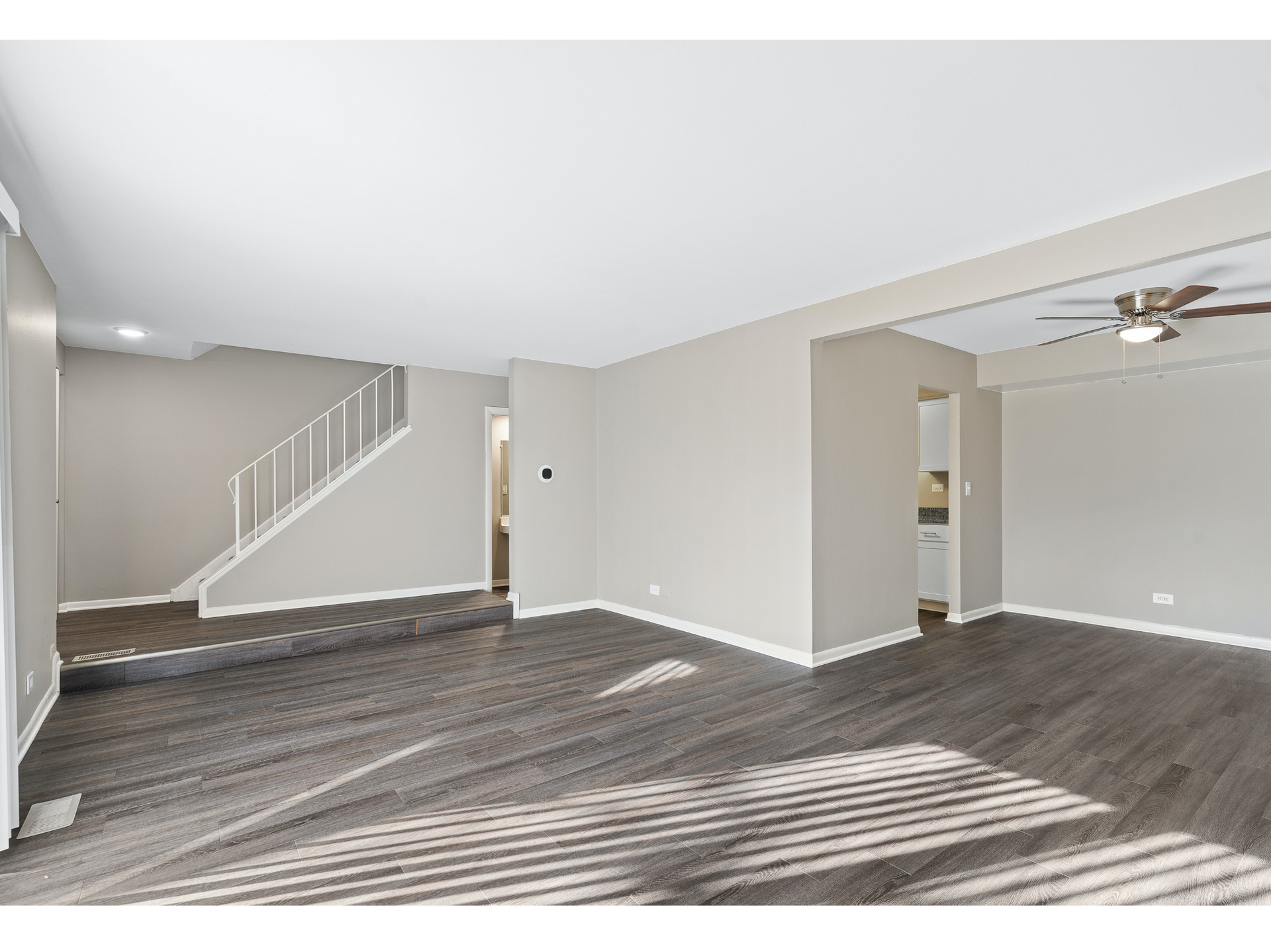 Empty living room with gray walls, carpeted floor, staircase, and adjacent dining area with ceiling fan