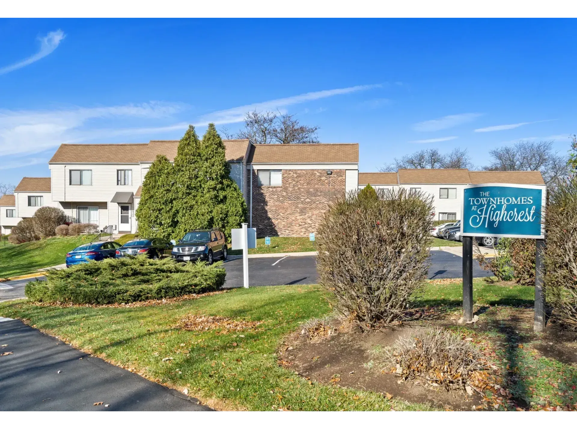 Apartment building with blue sign, parked cars, and shrubs on a sunny day