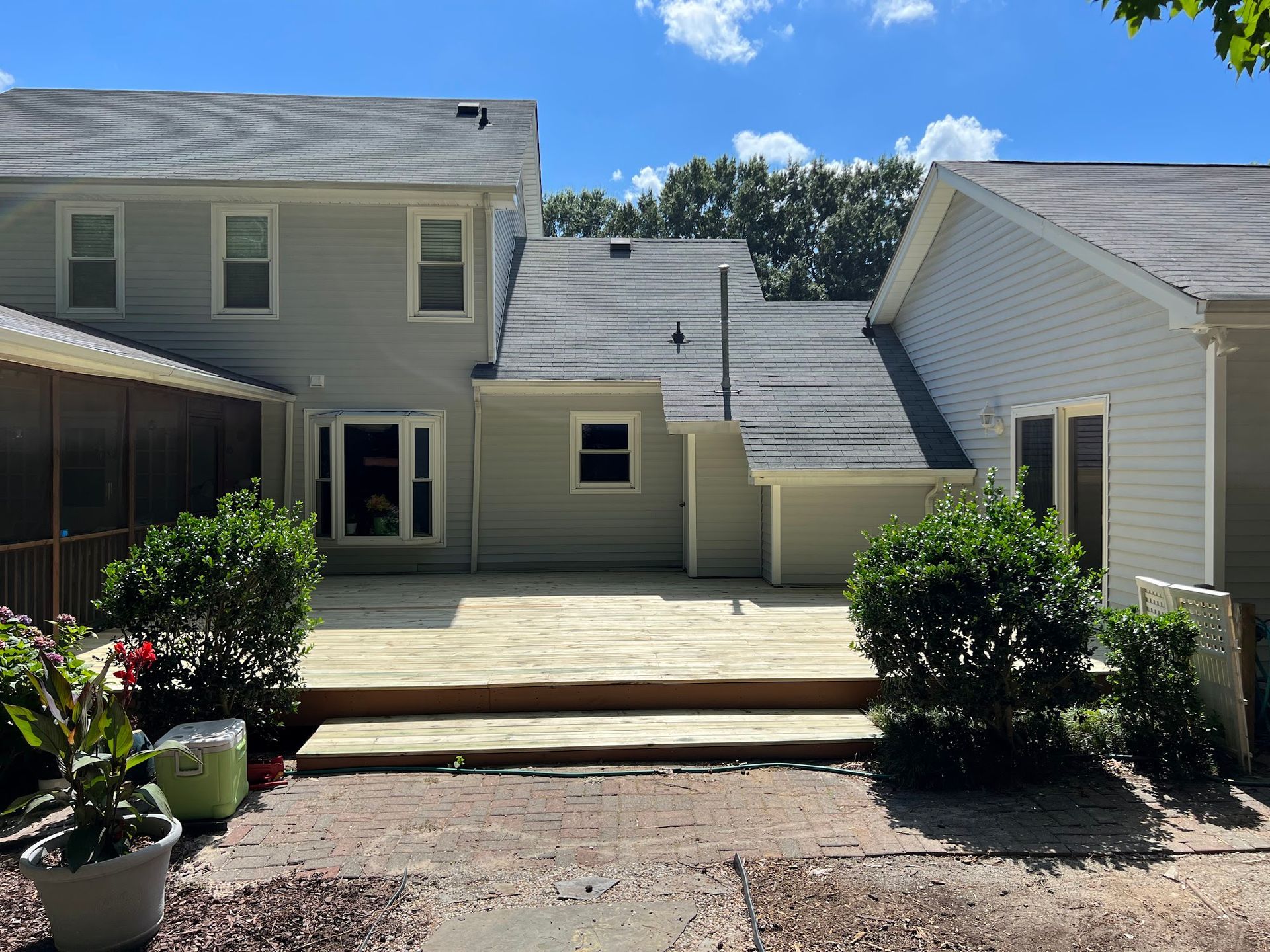 Backyard view of a two-story house with a wooden deck, plants, and a blue sky.