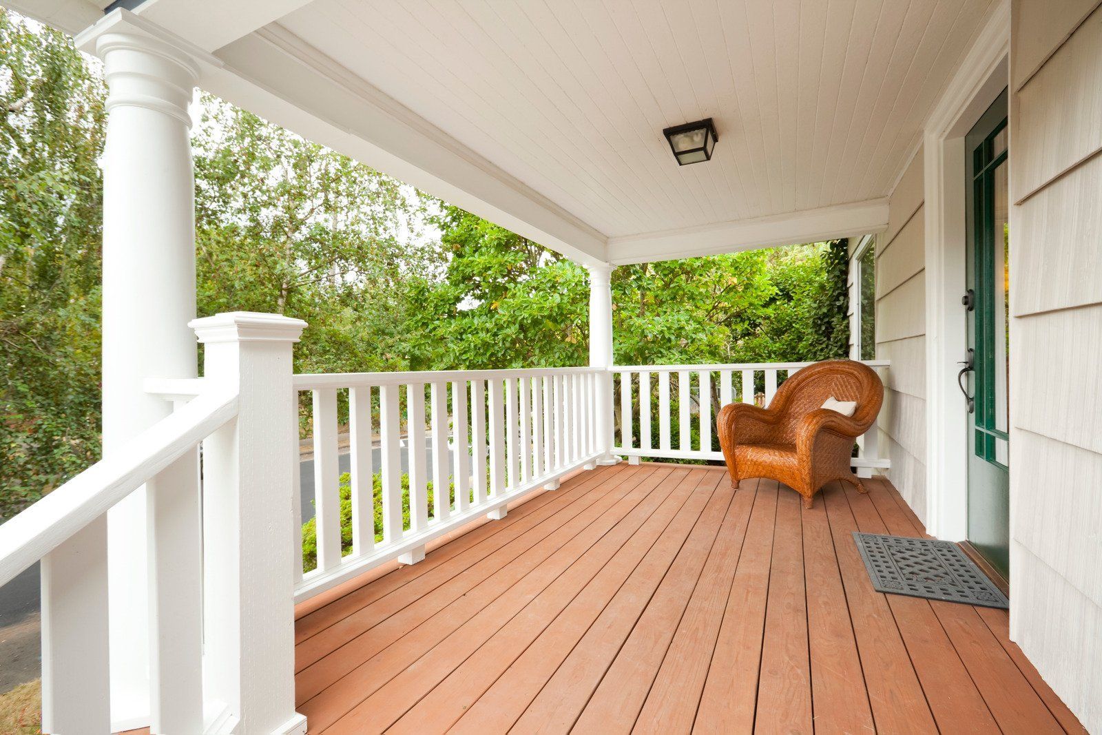 A porch with a brown wooden floor, white railings, and a wicker chair; green trees visible in the background.