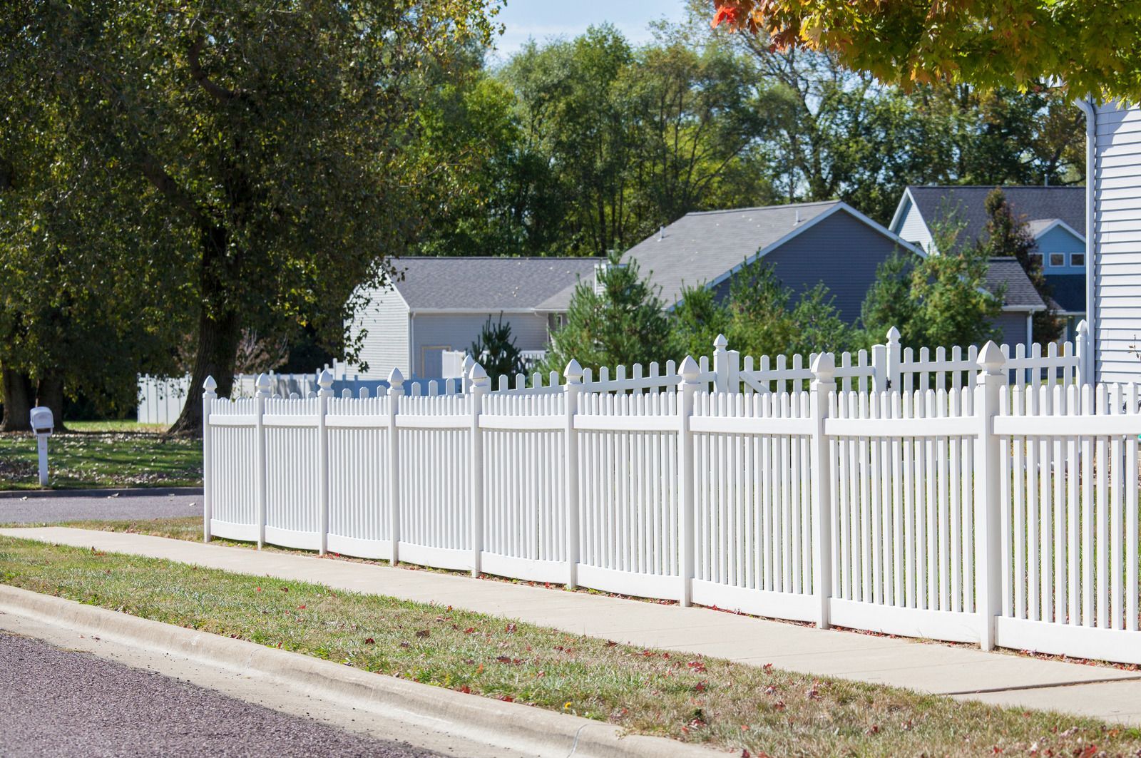 White picket fence bordering a sidewalk in front of houses, sunny day.