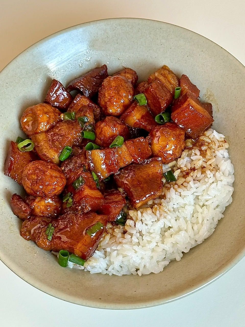 A beige bowl filled with braised pork belly, and white rice, garnished with green onions.