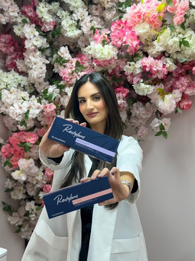 A woman is holding two boxes of injections in front of a wall of flowers.