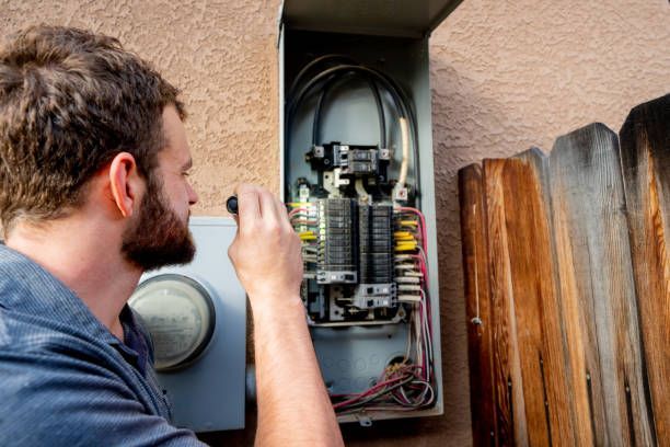 A person with a beard uses a small flashlight to inspect an open outdoor electrical panel.
