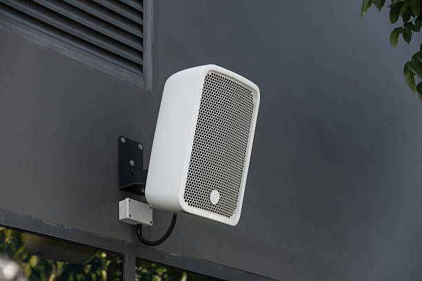 A white, rectangular outdoor loudspeaker mounted on a grey exterior wall.