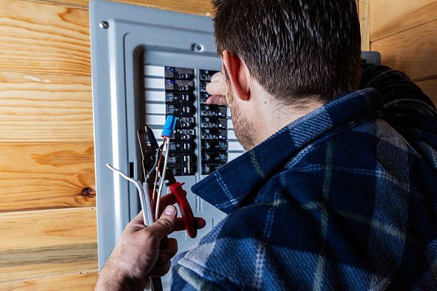 A person wearing a blue plaid shirt works on an open residential electrical breaker panel, holding wire strippers.