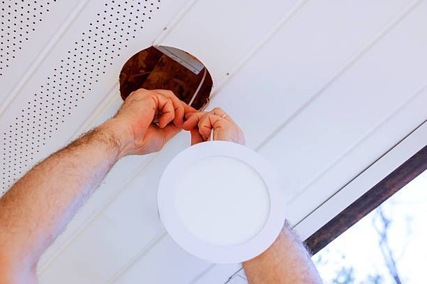 A person's hands installing a circular white recessed light fixture into a hole in a white soffit ceiling.