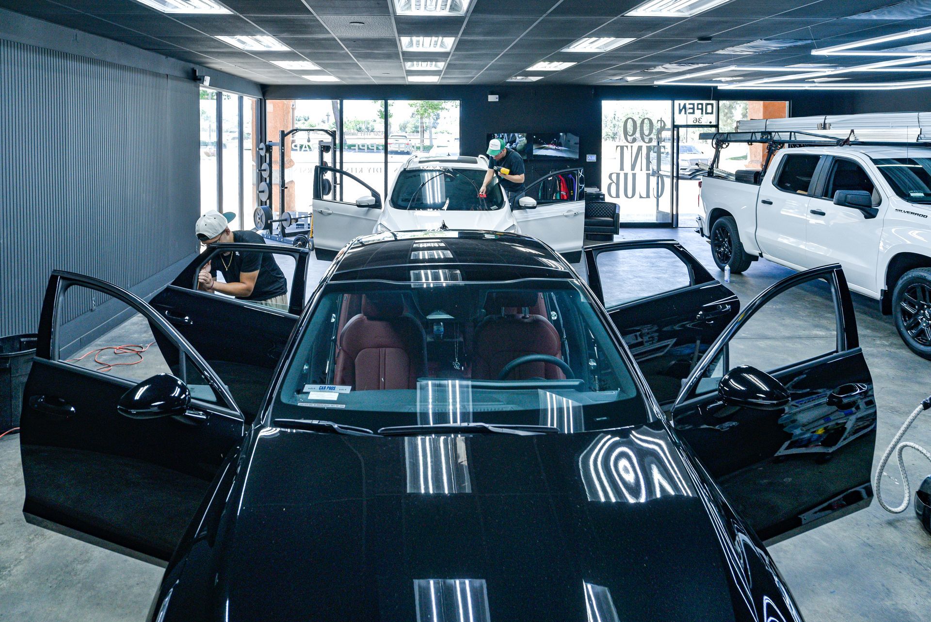 Black car with open doors at 99 Tint Club in Yucaipa, CA 's showroom, with white SUVs and a person tinting windows in the background
