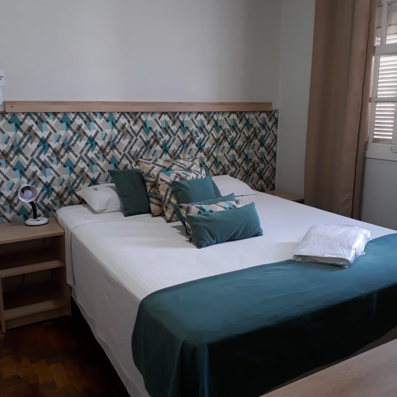 Um quarto estilo hotel com cama de lençóis brancos, cabeceira com estampa geométrica, almofadas decorativas em tom verde-azulado e móveis de madeira.