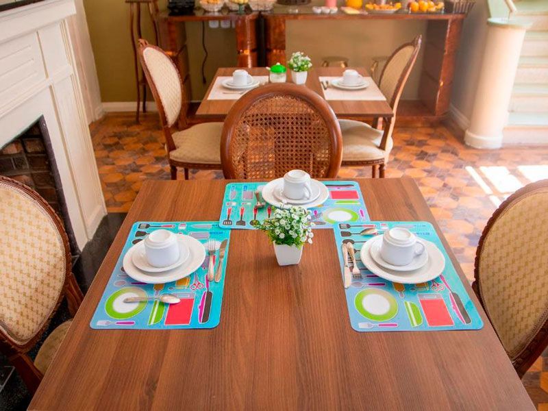 Uma mesa de jantar de madeira com quatro lugares e pequenos vasos de plantas, com cadeiras e outra mesa ao fundo.