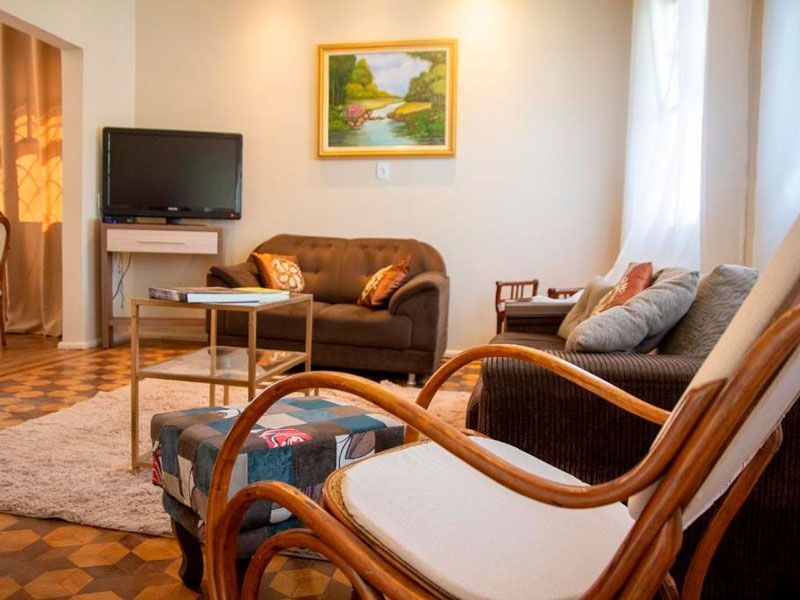 Uma sala de estar com sofás marrons, um pufe estampado, uma mesa de centro de vidro e uma TV sobre um suporte sob um quadro emoldurado.