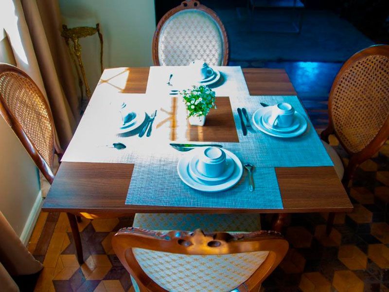 Uma mesa de jantar de madeira para quatro pessoas, posta com pratos e xícaras brancas, jogos americanos azuis e um pequeno arranjo de plantas verdes como centro de mesa.