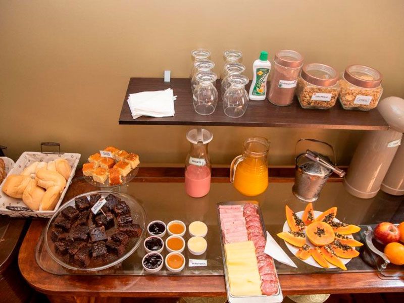 Um buffet de café da manhã com pães, brownies, doces, frios, queijos, mamão papaia, sucos, cereais e bebidas em copos.