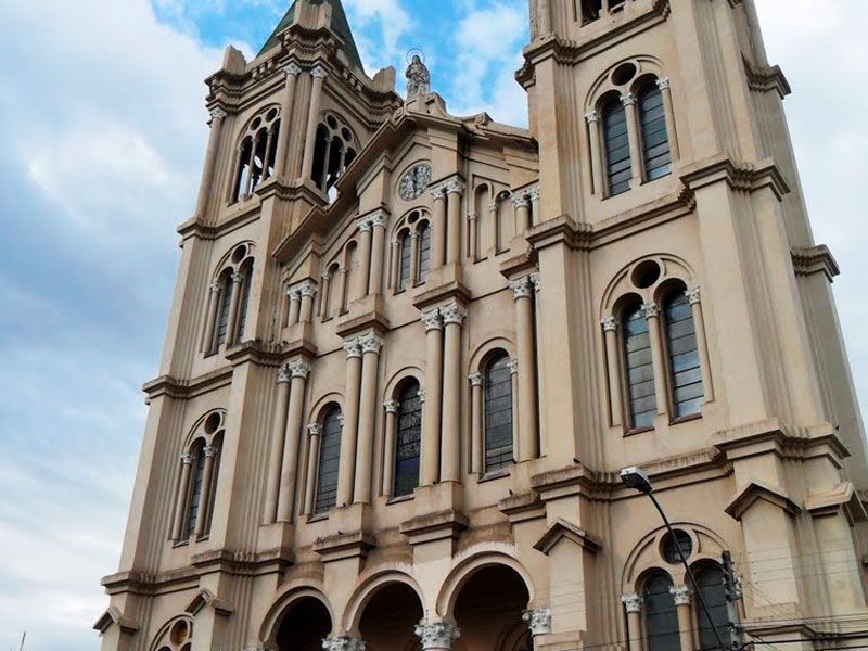 Vista de baixo para cima de uma catedral de pedra bege e ornamentada, com torres gêmeas e janelas em arco, sob um céu azul e nublado.