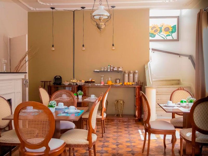 Sala de jantar com mesas e cadeiras, luminárias pendentes, um balcão de buffet encostado em uma parede bege e uma pintura de girassóis.