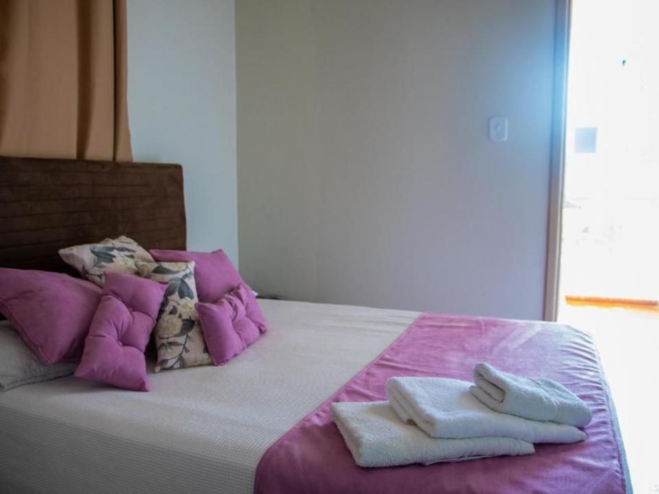 Uma cama com lençóis brancos, almofadas decorativas rosa, cabeceira marrom e toalhas brancas dobradas em um quarto iluminado.